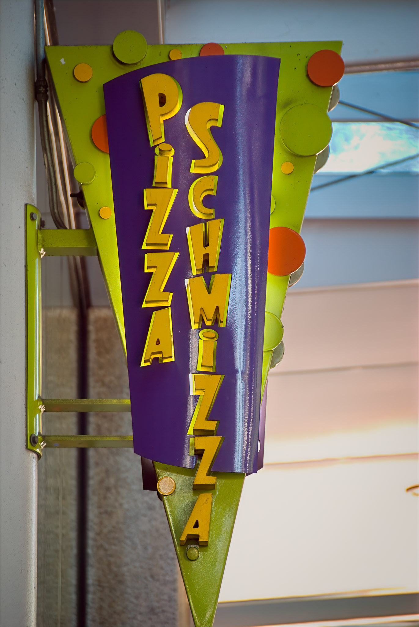 A colorful, triangular sign with the words Pizza Schmizza in playful, vertical yellow letters is displayed against a building exterior.