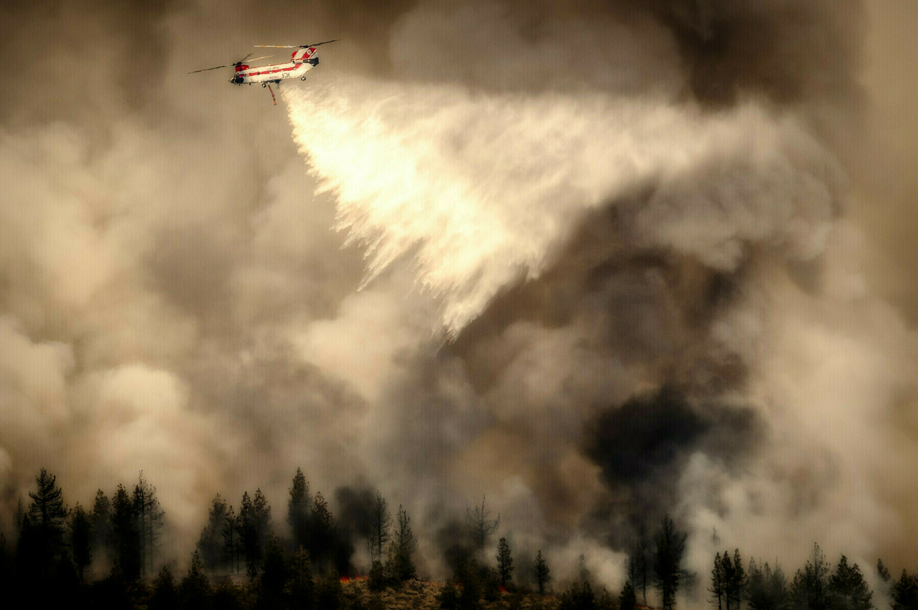 A helicopter drops water over a forest fire, creating a dramatic plume of smoke and mist.