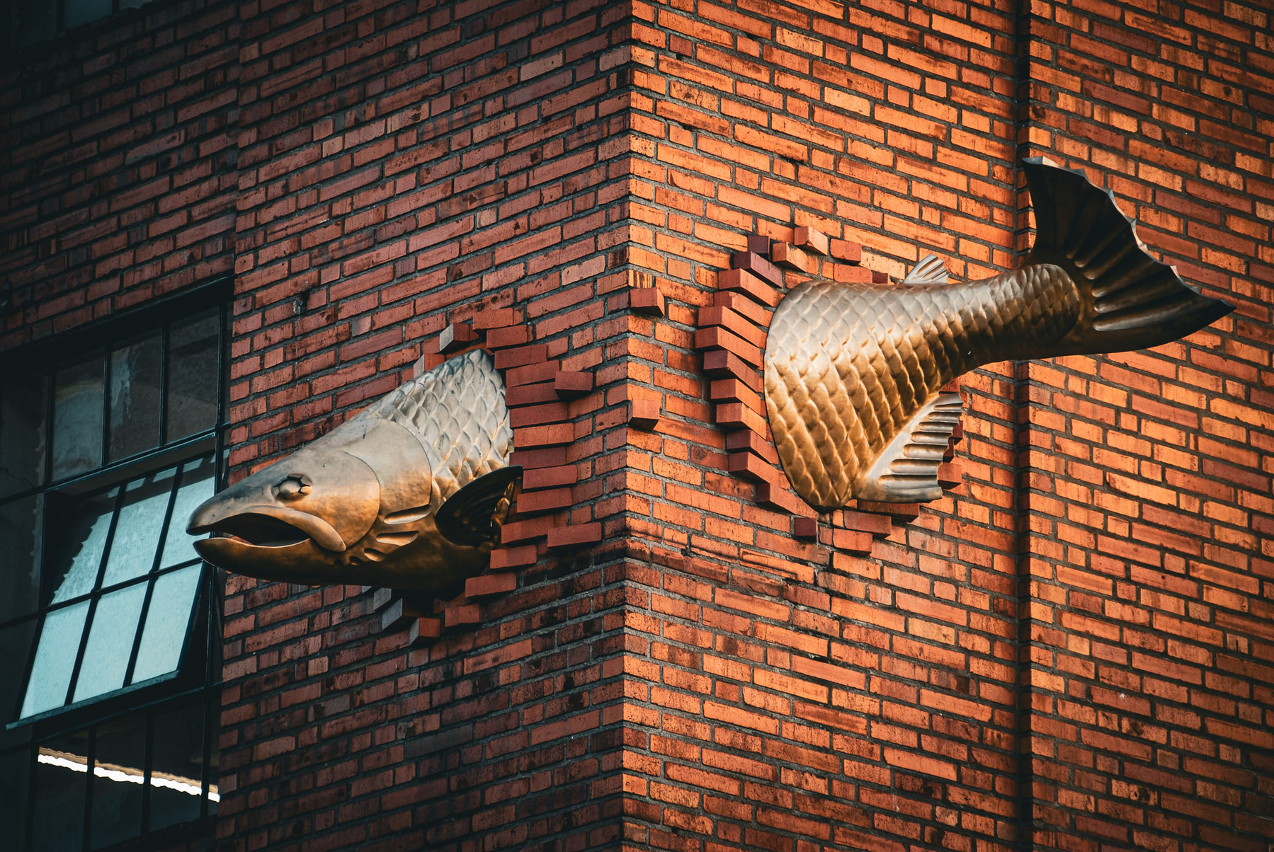 A sculpture of a large fish appears to be swimming through a brick wall, with its body on one side and tail on the other.