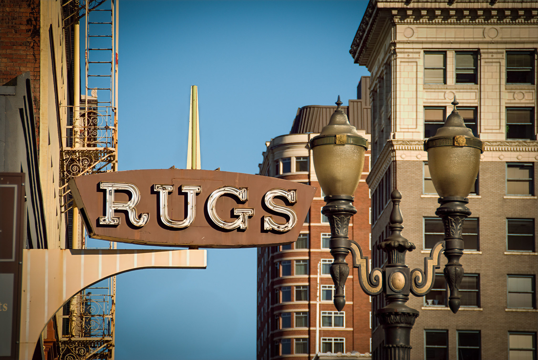 A vintage sign reading RUGS hangs on a city street with tall buildings and street lamps in the background.