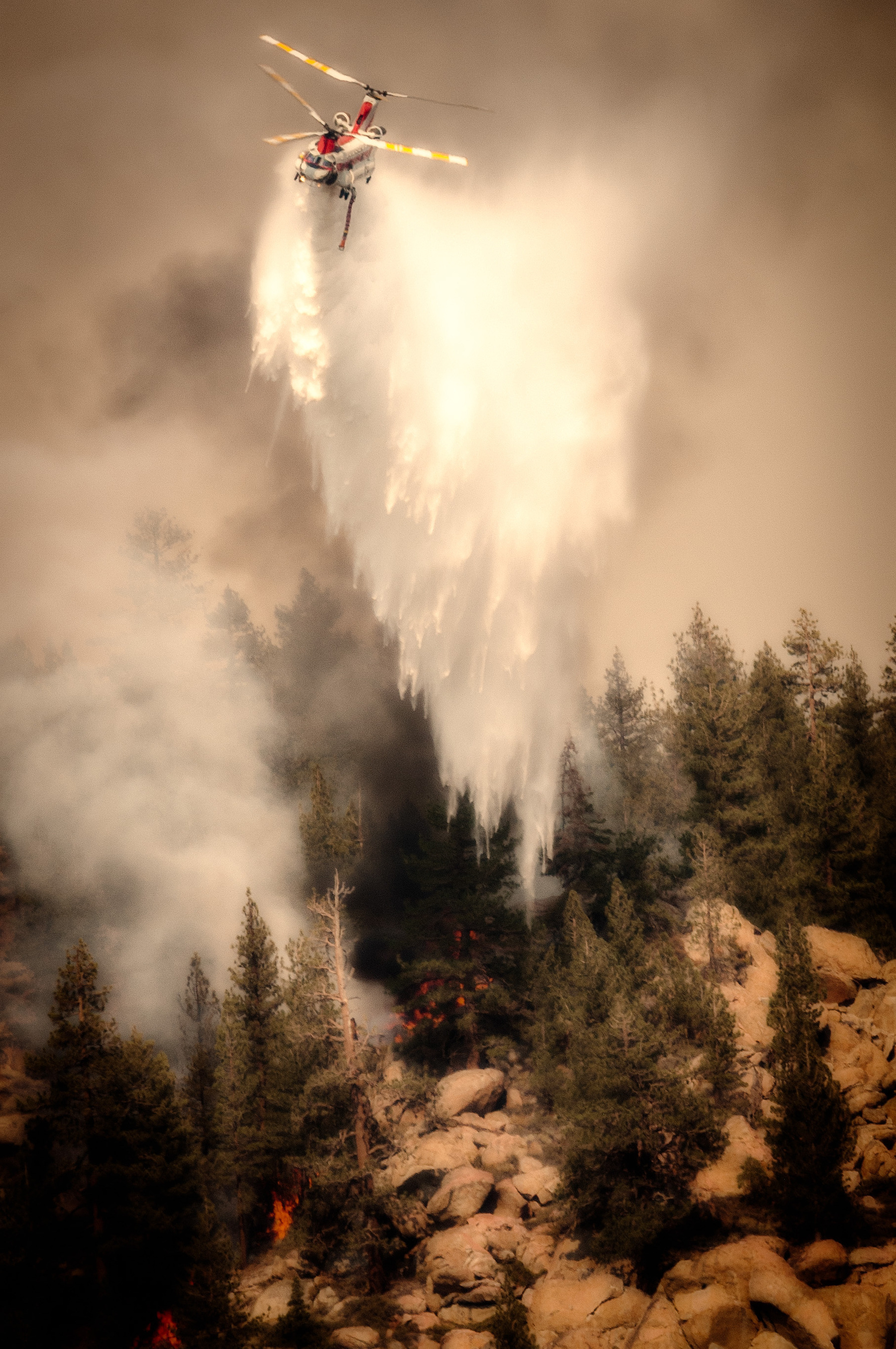 Auto-generated description: A helicopter is dropping water over a forest fire to extinguish the flames.