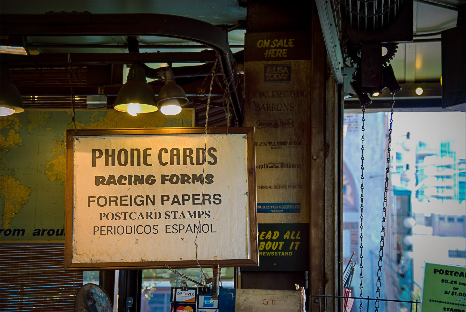 A sign in a shop advertises phone cards, racing forms, foreign papers, postcards, stamps, and periodicals in Spanish.