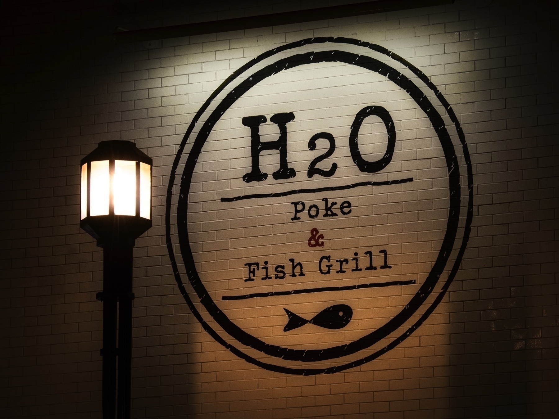 Auto-generated description: A round sign for H2O Poke & Fish Grill is illuminated on a brick wall next to a street lamp.