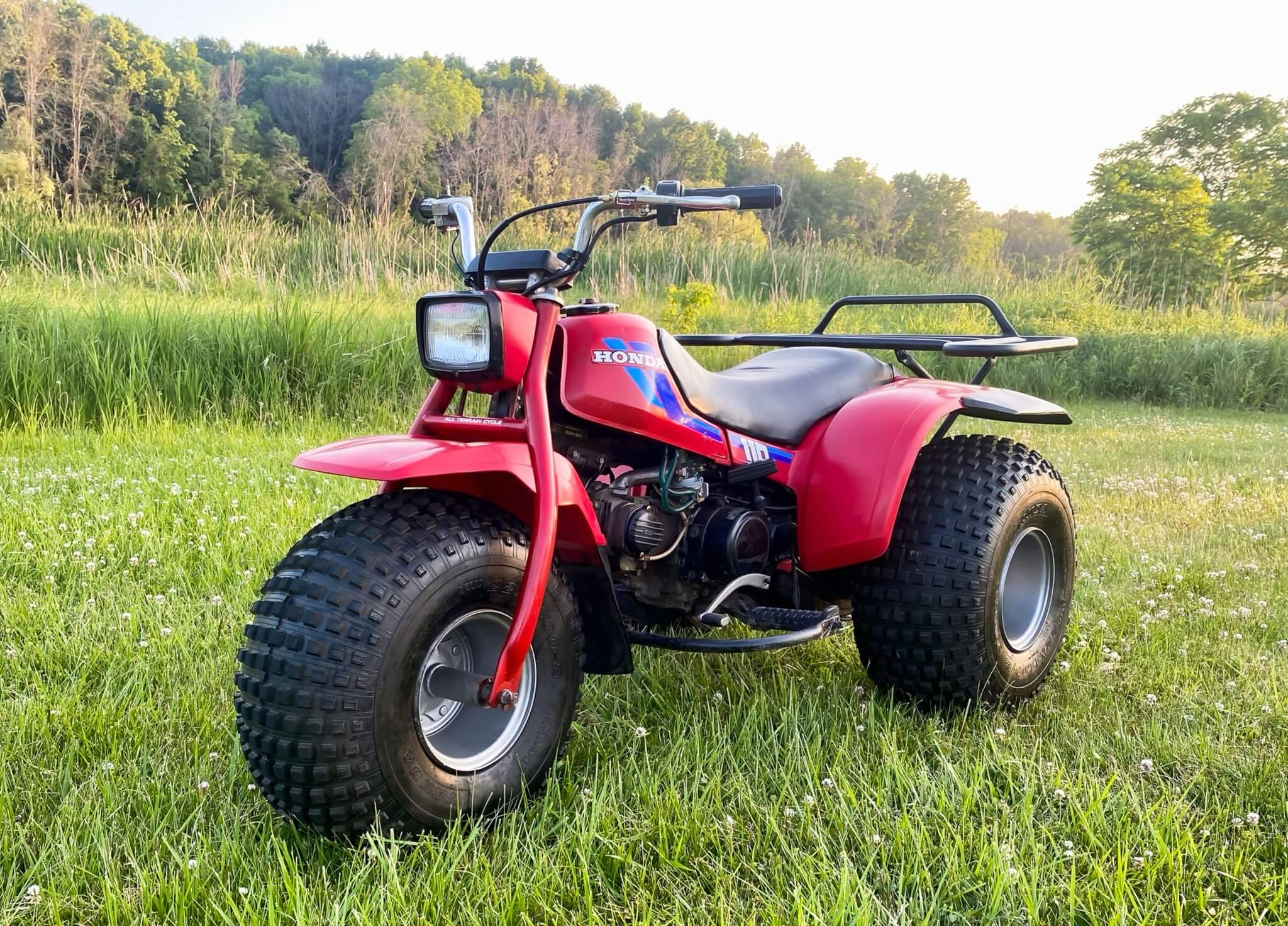 Auto-generated description: A red three-wheeler ATV is parked on a grassy field with a tree-lined background.