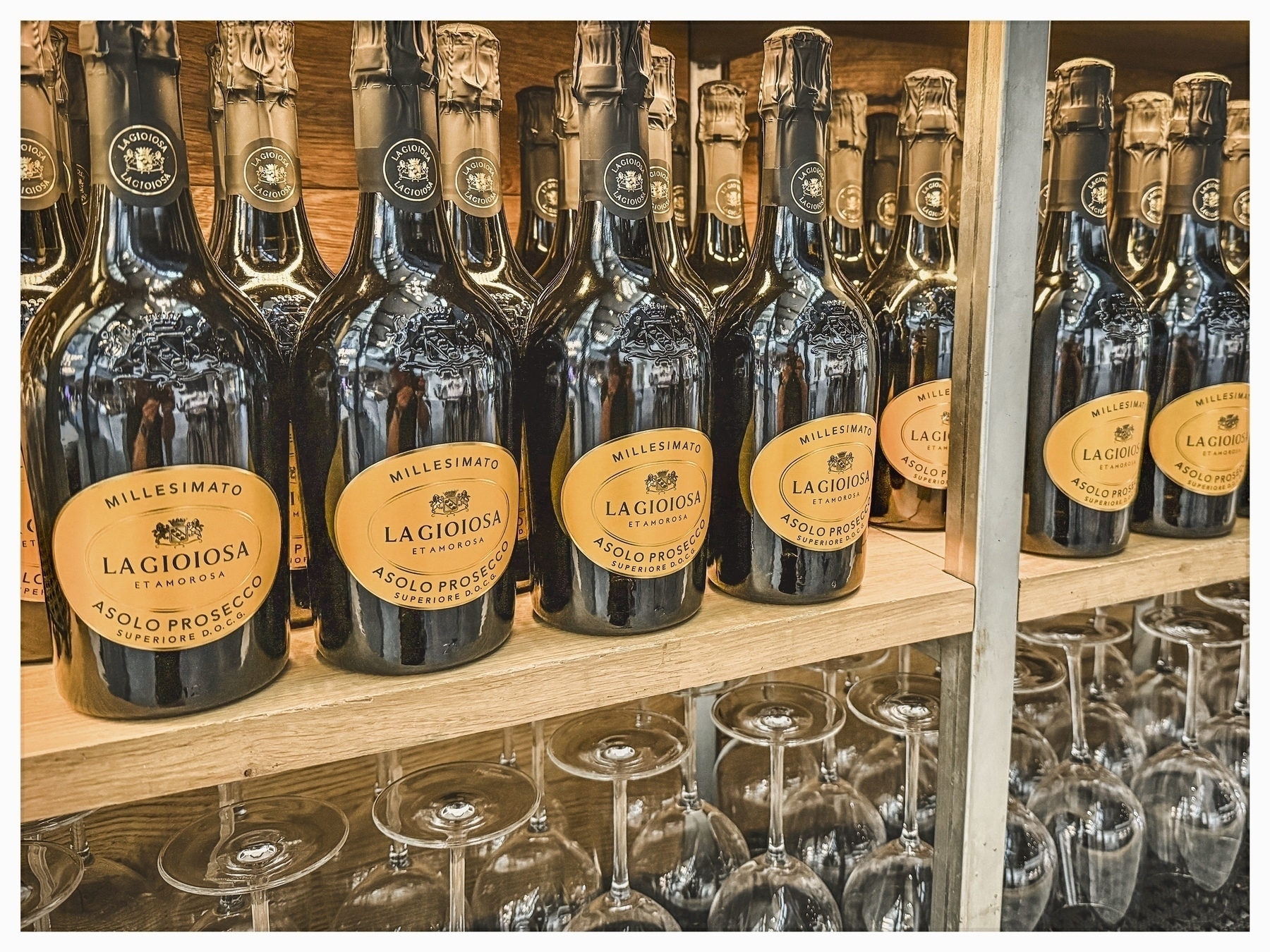 Auto-generated description: Several bottles of La Gioiosa Asolo Prosecco are displayed on wooden shelves with rows of upside-down wine glasses beneath them.