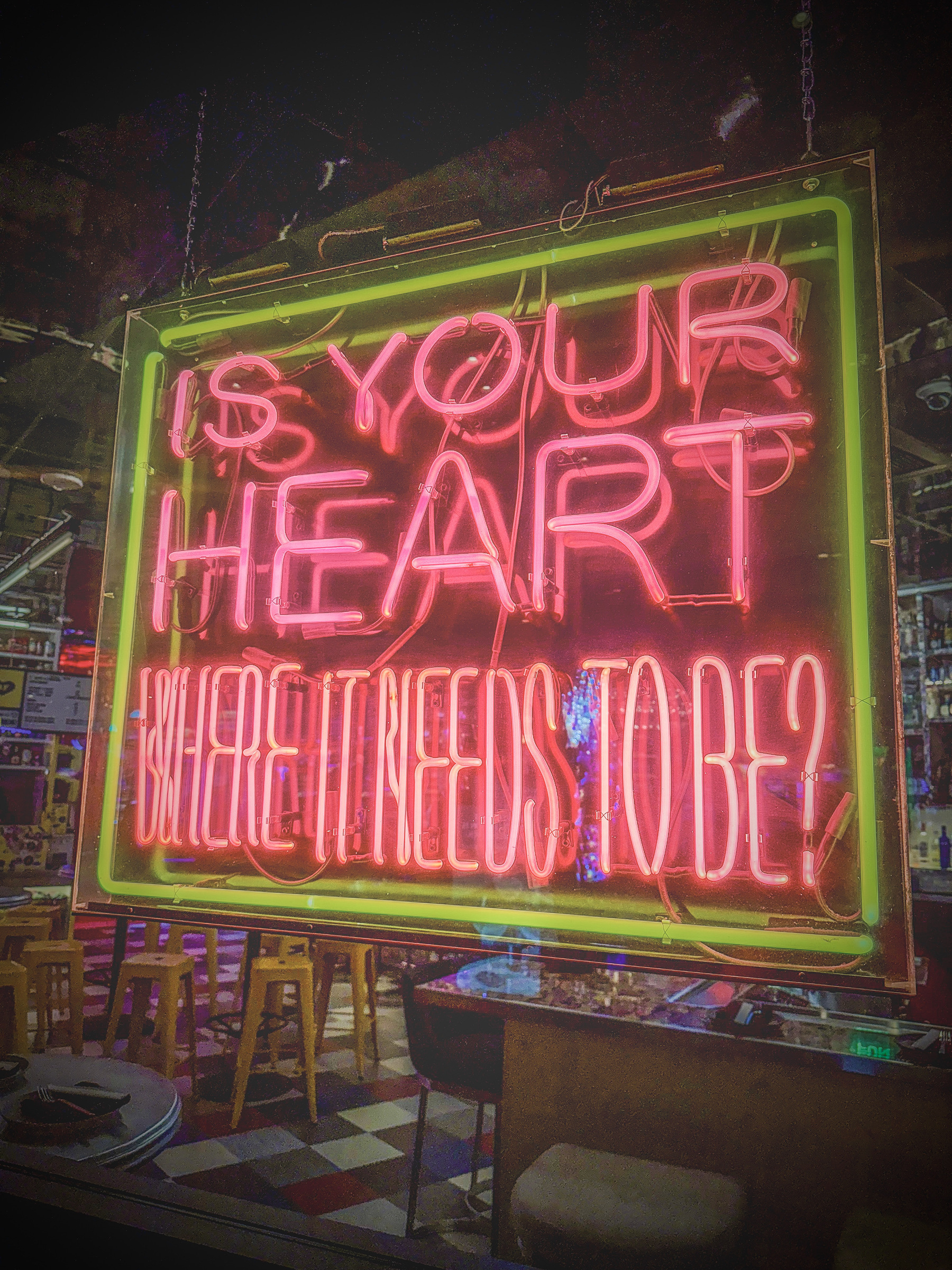 Auto-generated description: A neon sign in a bar or restaurant displays the phrase IS YOUR HEART WHERE IT NEEDS TO BE?.