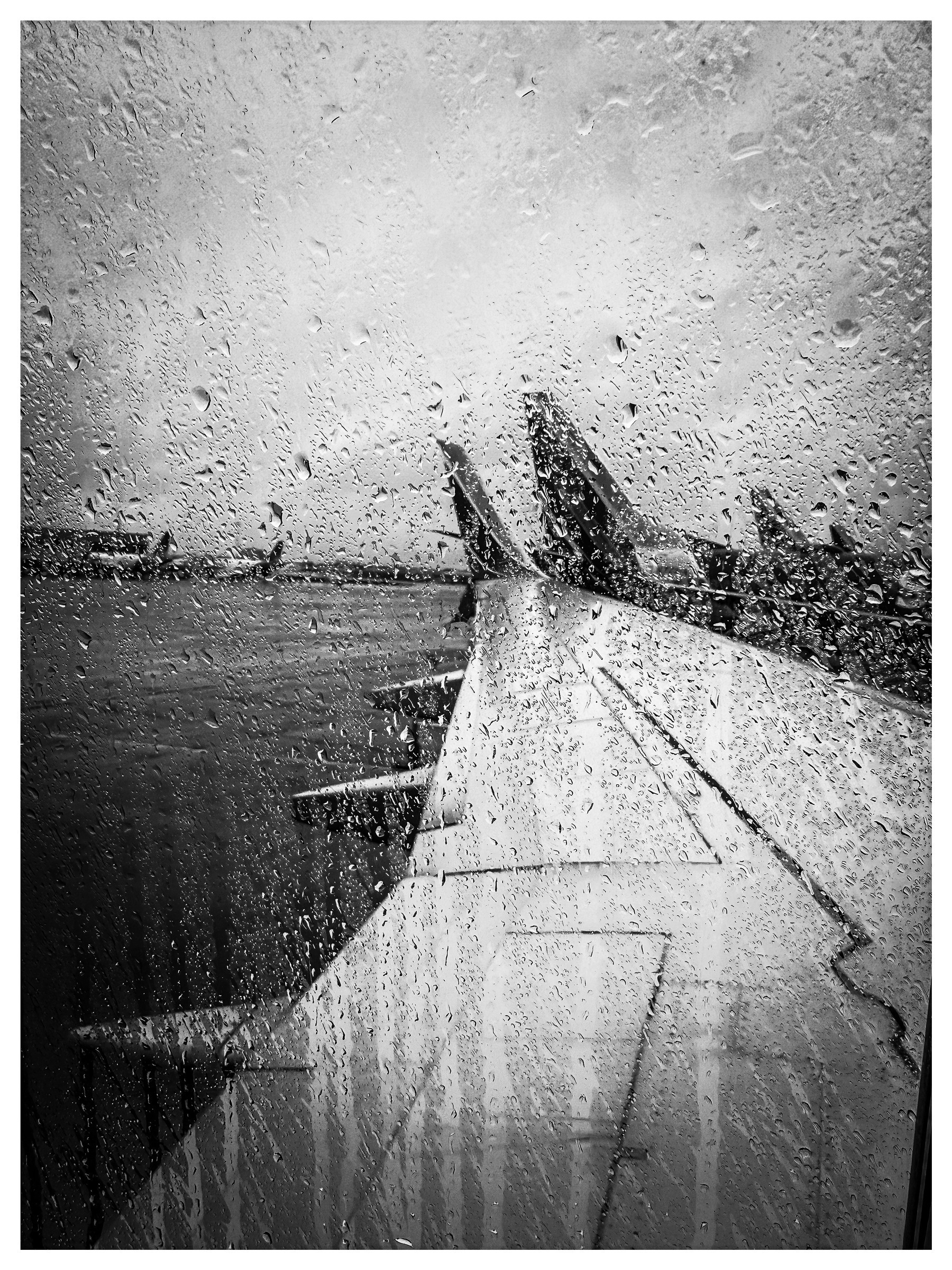 Auto-generated description: Raindrops cover an airplane window, offering a view of the wing and several planes on a cloudy day at an airport.