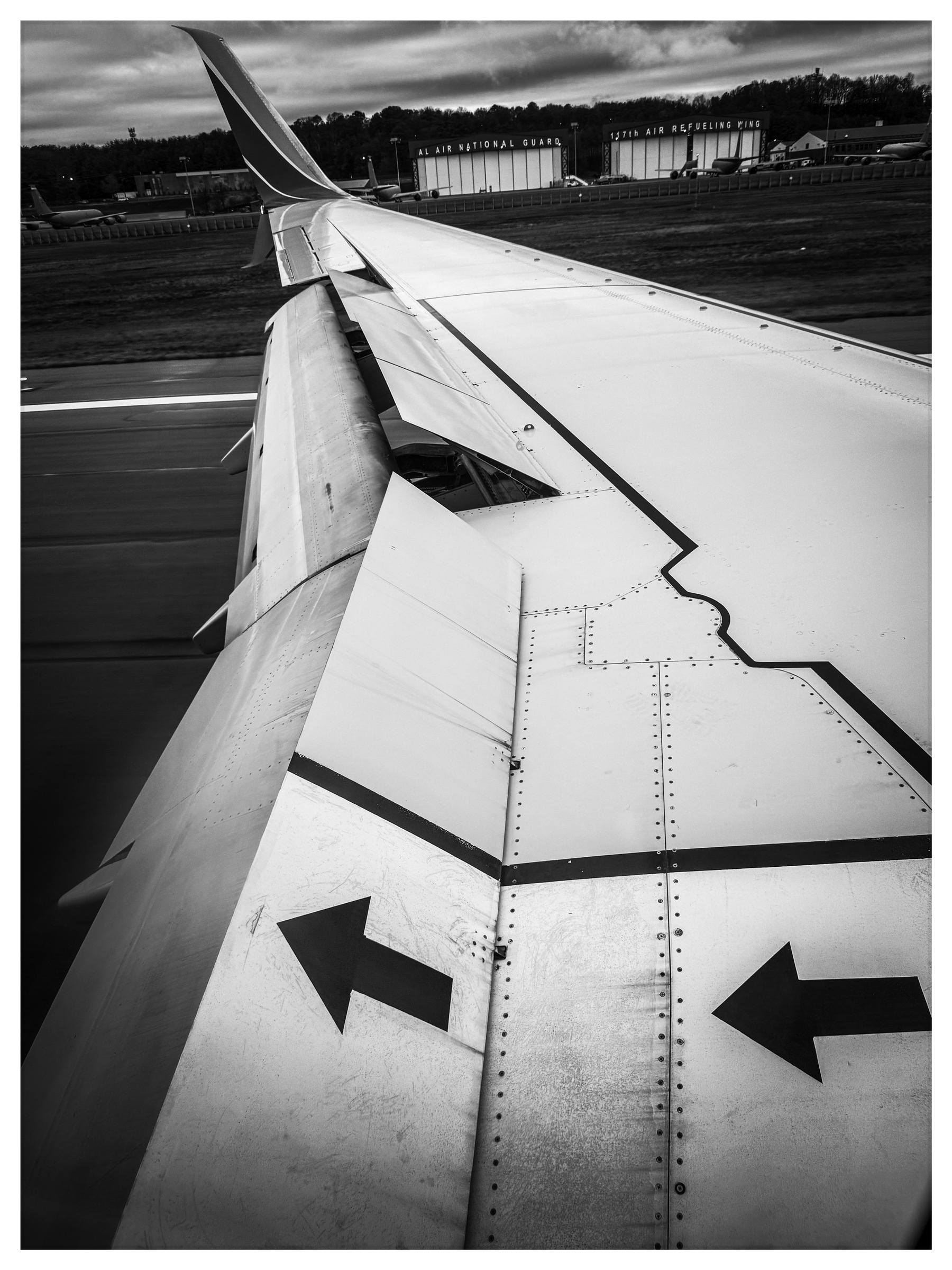 Auto-generated description: A black and white view from an airplane window showcases the wing with visible flaps and directional arrows.