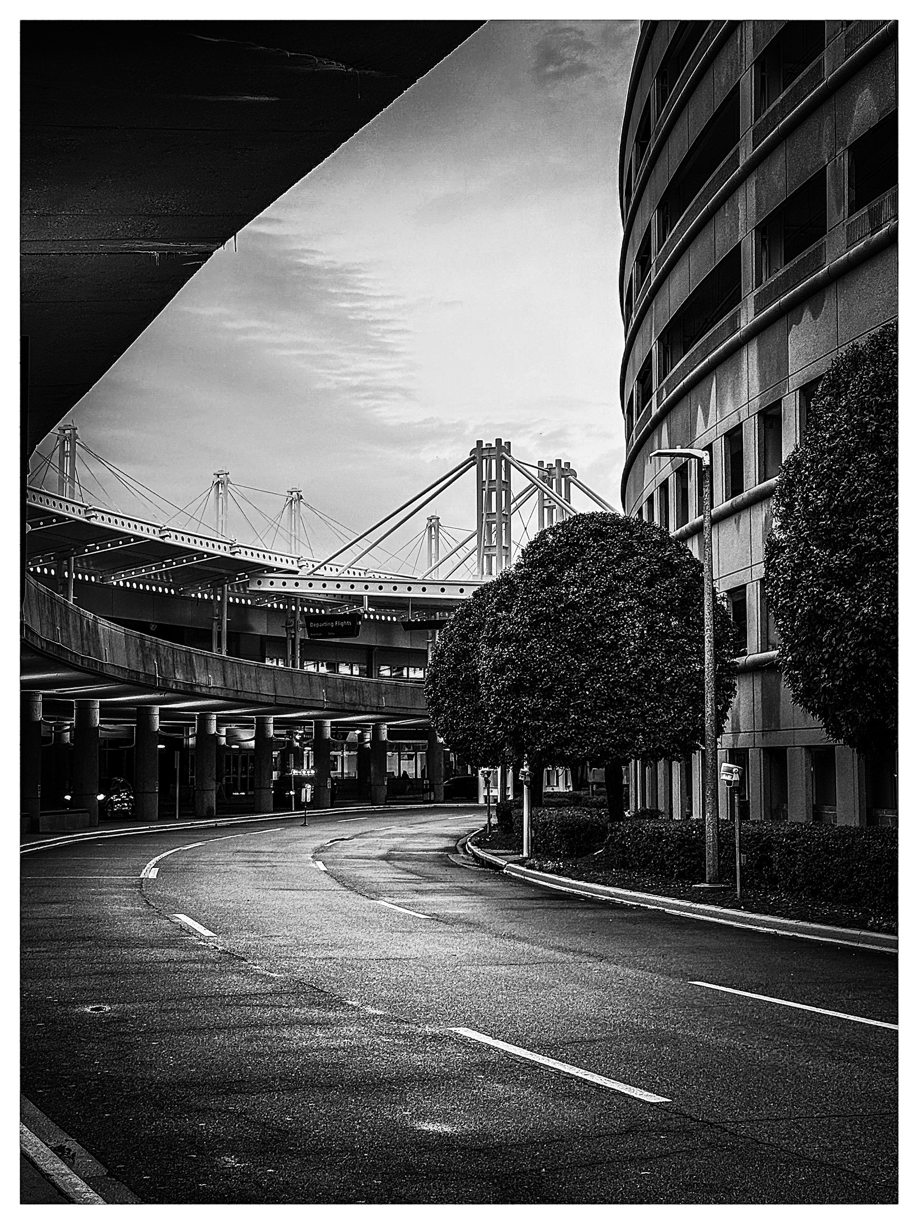 Auto-generated description: A curved road lined with trees passes by modern rounded buildings against the backdrop of a suspension bridge under a moody sky.