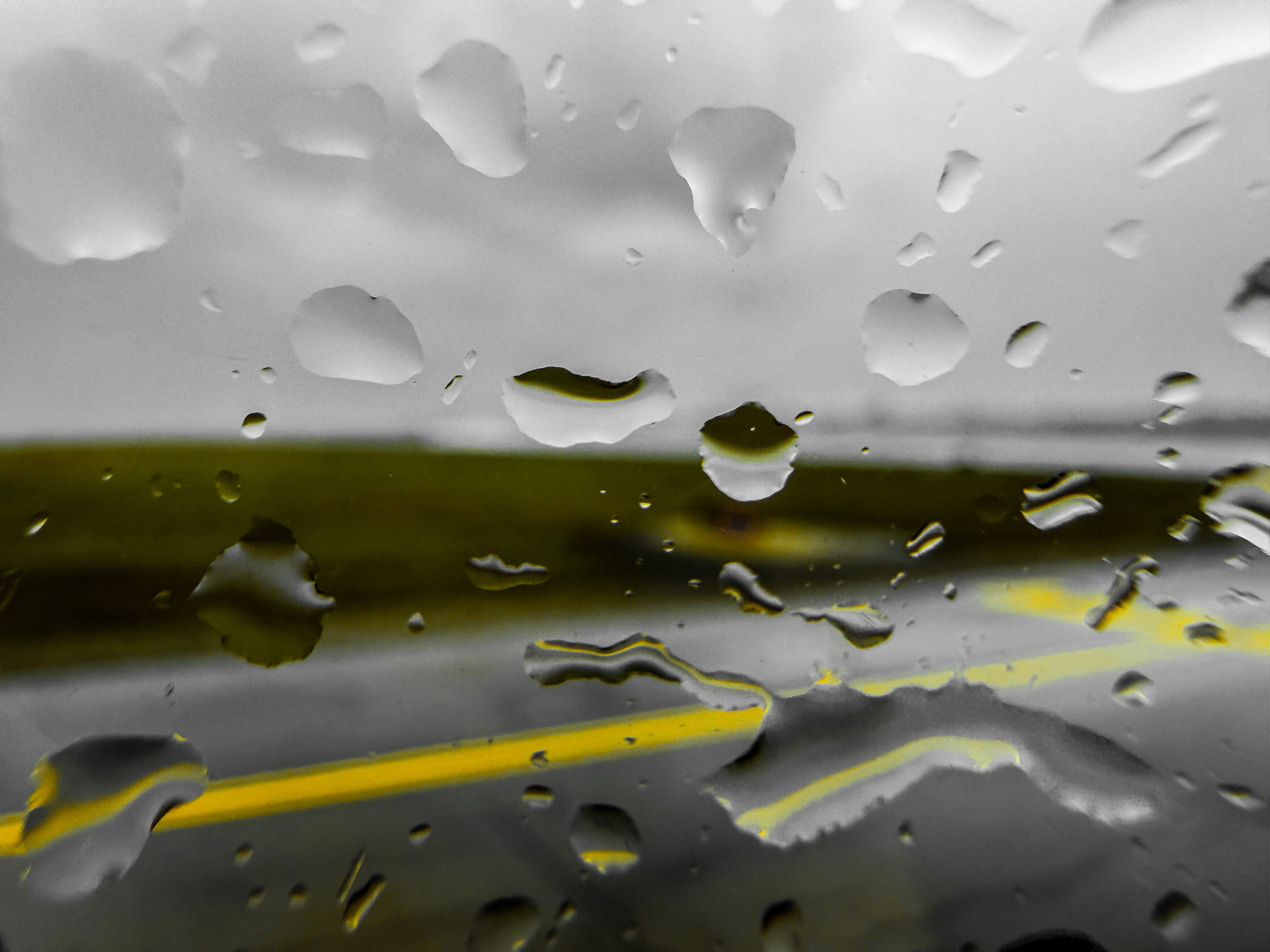 Auto-generated description: Raindrops are scattered on a window, with blurred views of a road and grassy landscape in the background.