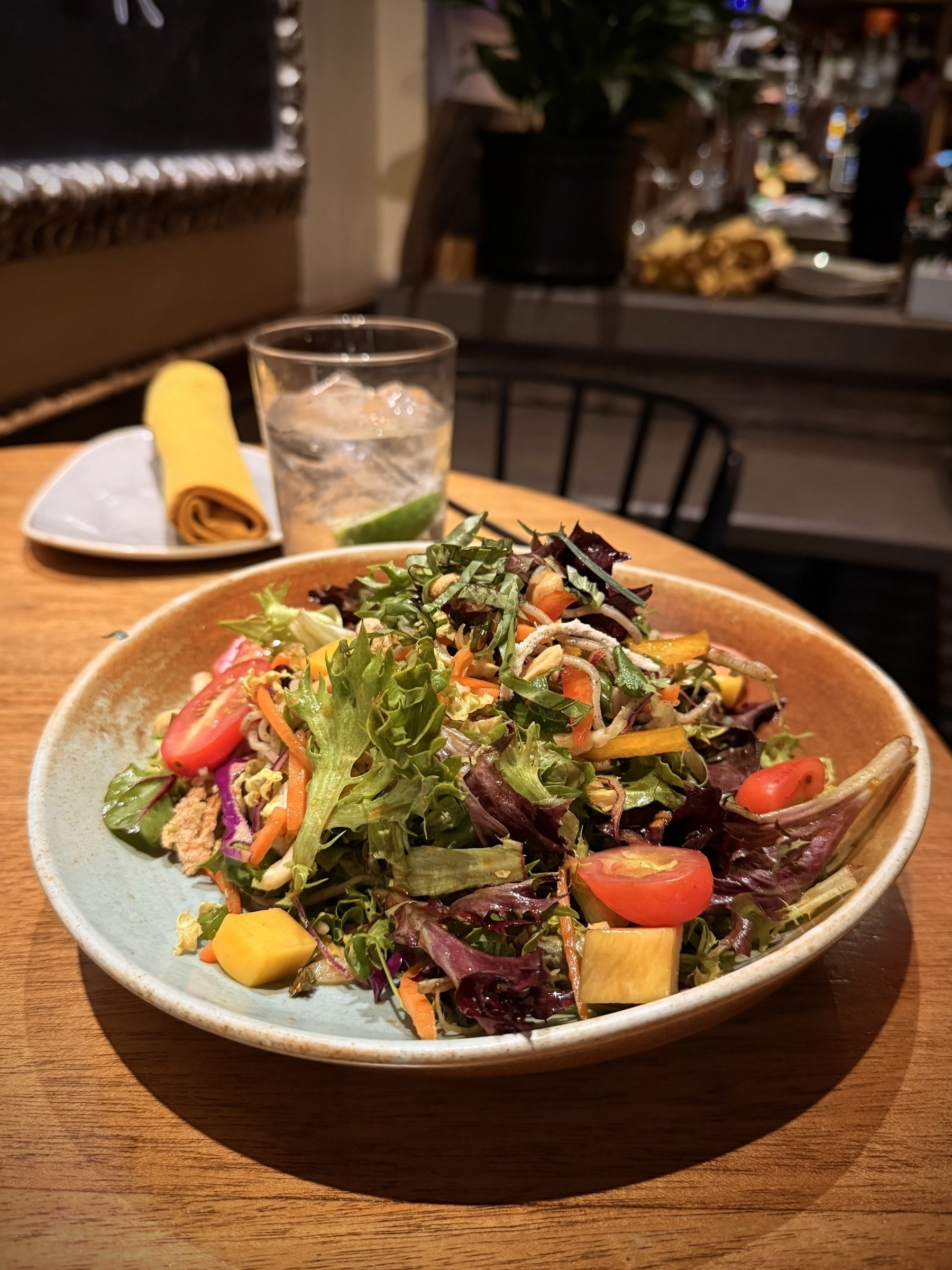 Auto-generated description: A colorful salad with assorted greens, cherry tomatoes, and mango pieces is served on a ceramic plate alongside a glass of water and a napkin on a wooden table.