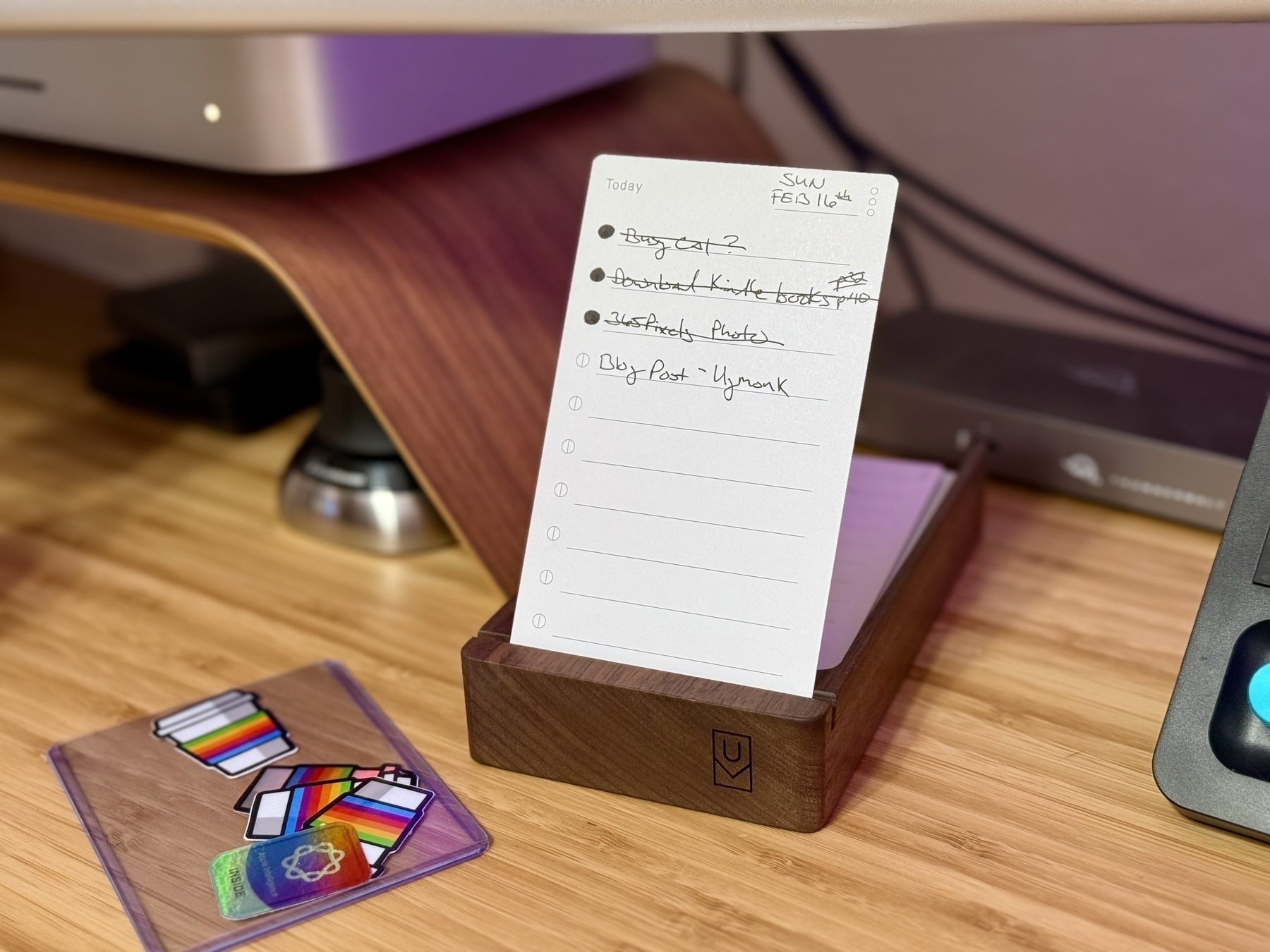 Auto-generated description: A to-do list placed on a wooden holder sits on a desk near a computer, with a small card featuring colorful icons nearby.