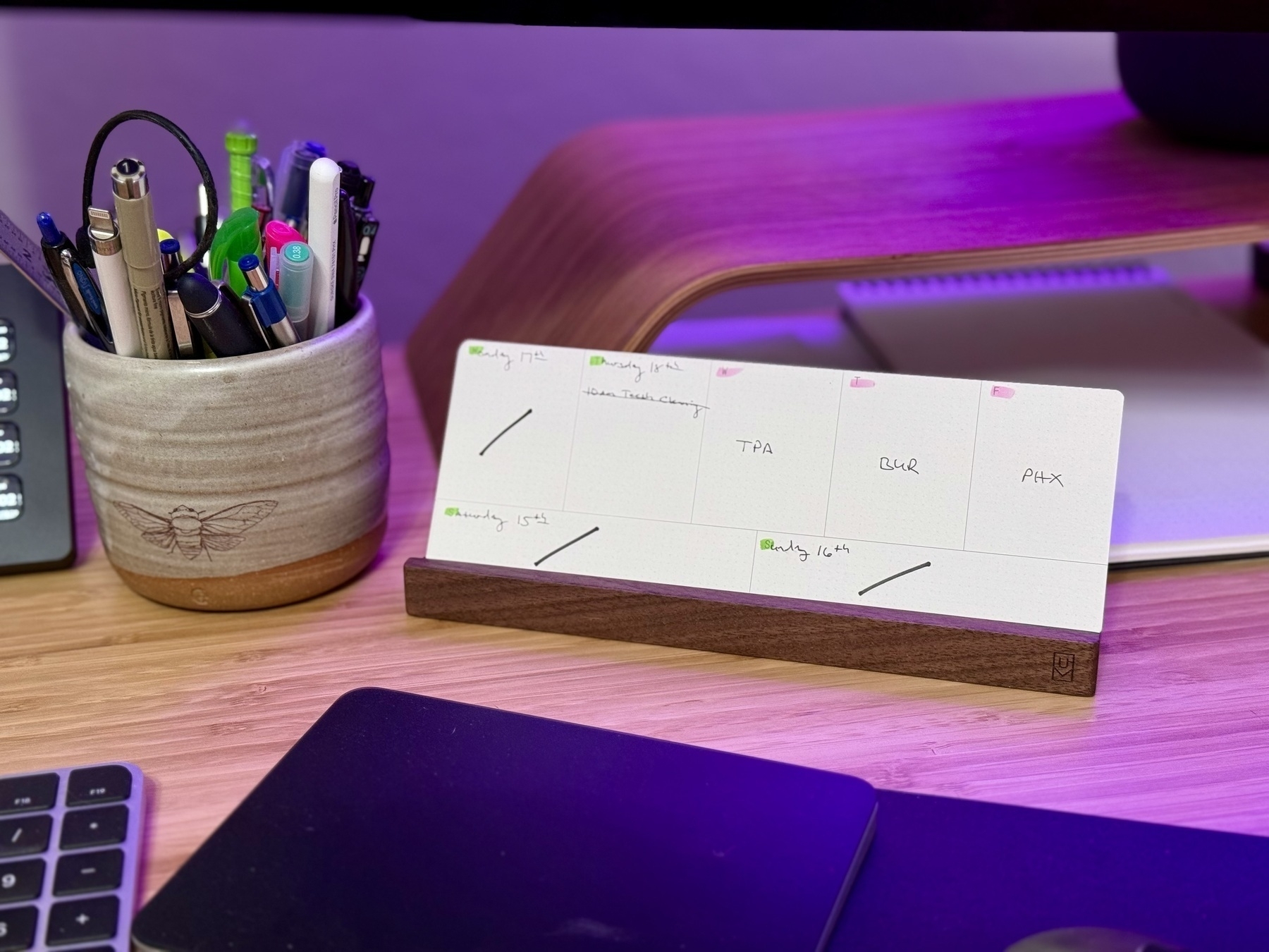 Auto-generated description: A desk features a container of pens and markers, an open weekly planner in a wooden holder, a portion of a keyboard, and a mouse pad.