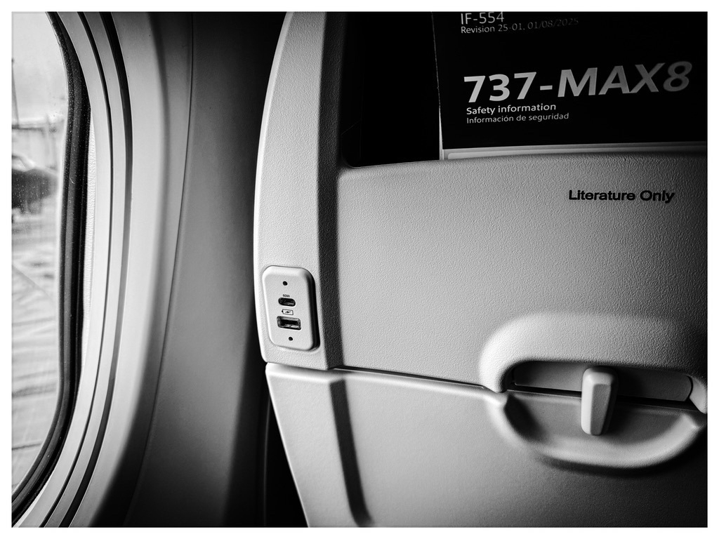 Auto-generated description: A black and white view of an airplane window seat area, featuring a safety information card for a 737-MAX8 and a tray table marked Literature Only.