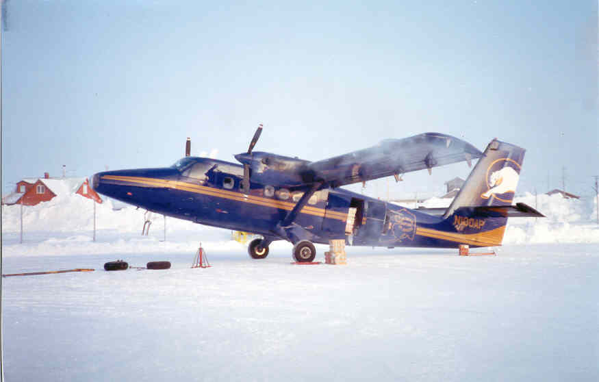 Auto-generated description: A blue and yellow airplane is parked on a snowy surface surrounded by ice and a few scattered items, with red-roofed buildings in the background.