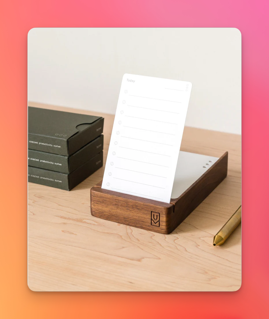 Auto-generated description: A wooden stand holds a notepad with a list template, placed on a wooden surface next to a pen and stacked boxes.