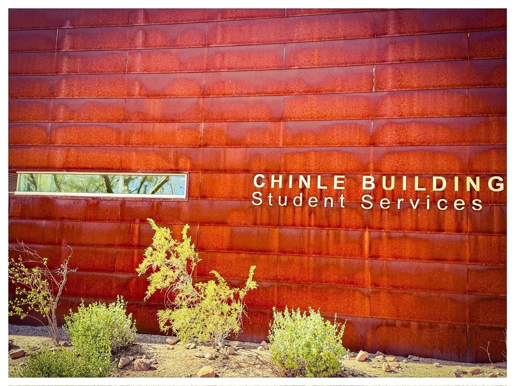 Auto-generated description: A rust-colored wall labeled Chinle Building Student Services is accompanied by small desert plants in the foreground.