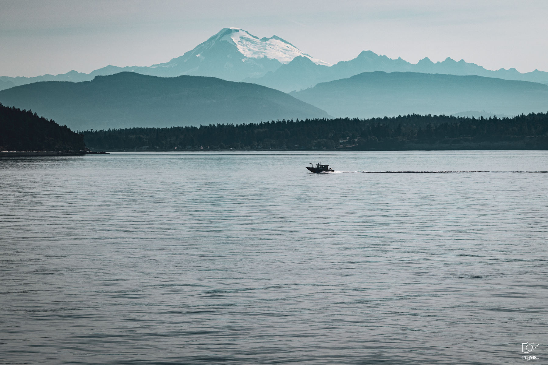 Auto-generated description: A boat is cruising on a calm body of water with snow-capped mountains and forested hills in the background under a clear sky.